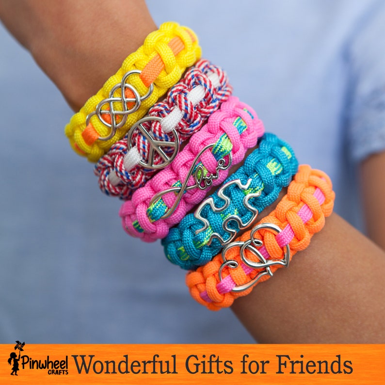 May include: A stack of five colorful friendship bracelets with different charms, including an infinity symbol, a peace sign, and a heart. The bracelets are made of woven paracord and are a great gift for friends.