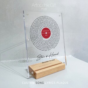 May include: A clear acrylic plaque with a circular design featuring song lyrics from the song "It's All About You" by McFly. The plaque is on a wooden stand and has the names "Steve & Hannah" and the date "December 3rd 2022" inscribed on it. The text "CUSTOM SONG LYRICS PLAQUE" is printed below the stand.