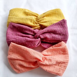 May include: Three fabric headbands in different colors: yellow, pink, and orange. Each headband has a knotted design and is covered in small gold glitter dots.