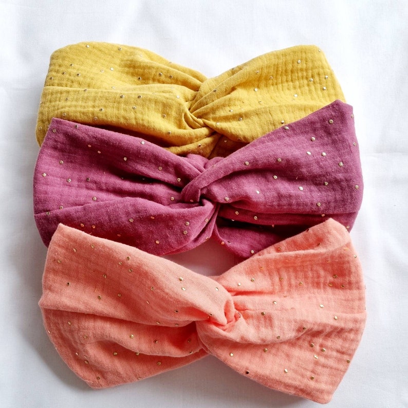 May include: Three fabric headbands in different colors: yellow, pink, and orange. Each headband has a knotted design and is covered in small gold glitter dots.