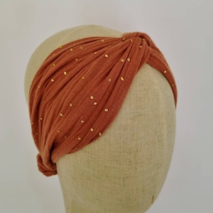 May include: A rust-colored turban headband with gold glitter accents. The headband is made of a soft, stretchy fabric and is designed to be worn as a knotted turban.