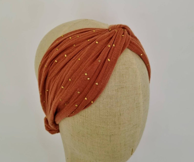 May include: A rust-colored turban headband with gold glitter accents. The headband is made of a soft, stretchy fabric and is designed to be worn as a knotted turban.