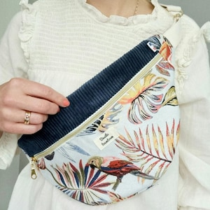 Puede incluir: Una riñonera blanca con una solapa de pana azul y cierre de cremallera. La parte delantera de la riñonera presenta un estampado tropical colorido con loros y hojas de palmera. El texto "Basile Tourdumonde" está impreso en la parte delantera de la riñonera.