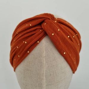 May include: A rust-colored turban headband with gold glitter dots. The headband is made of a soft, stretchy fabric and is designed to be worn as a fashion accessory.
