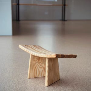 May include: A wooden meditation stool with a curved top and two legs. The stool is made of light brown wood and has a natural finish.