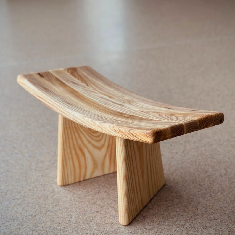 May include: A wooden meditation stool with a curved top and two legs. The stool is made of light brown wood with a natural finish.