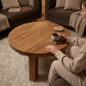 May include: A round wooden coffee table with a natural wood grain finish. The table has a single, thick leg and is surrounded by a cream-colored rug. A person is holding a cup of tea on the table. The table is approximately 36 inches in diameter.