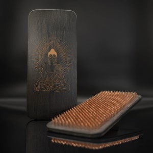 May include: A black wooden board with a gold-colored image of a meditating Buddha in a triangle with rays of light. A black wooden board with copper-colored pins is in the foreground.