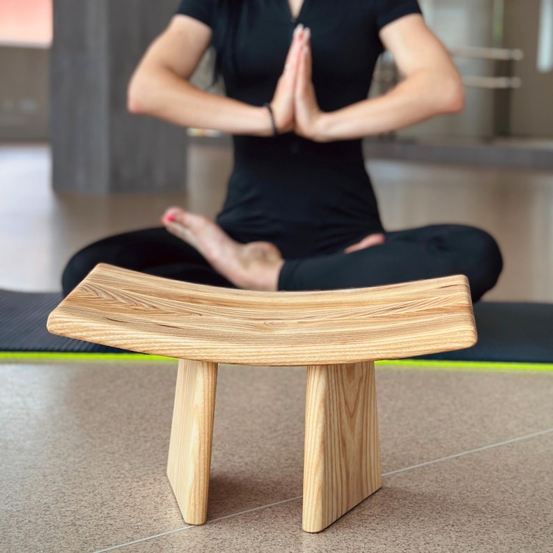 May include: A wooden meditation cushion with a curved top and three legs. The cushion is made of light brown wood and has a natural finish.