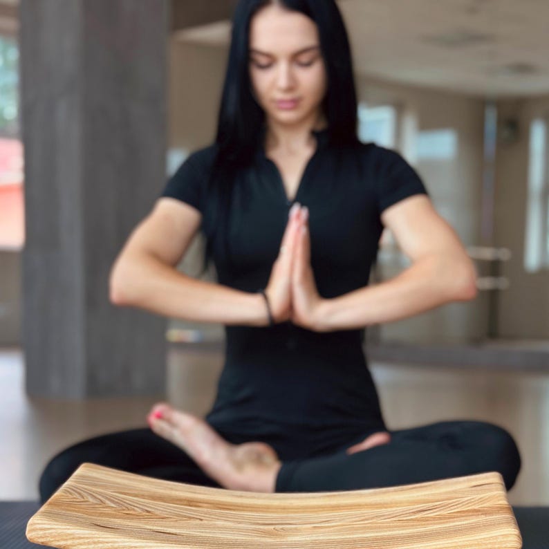 May include: A wooden meditation cushion with a curved shape and a natural wood grain finish. The cushion is in focus, while a person in the background is blurred.