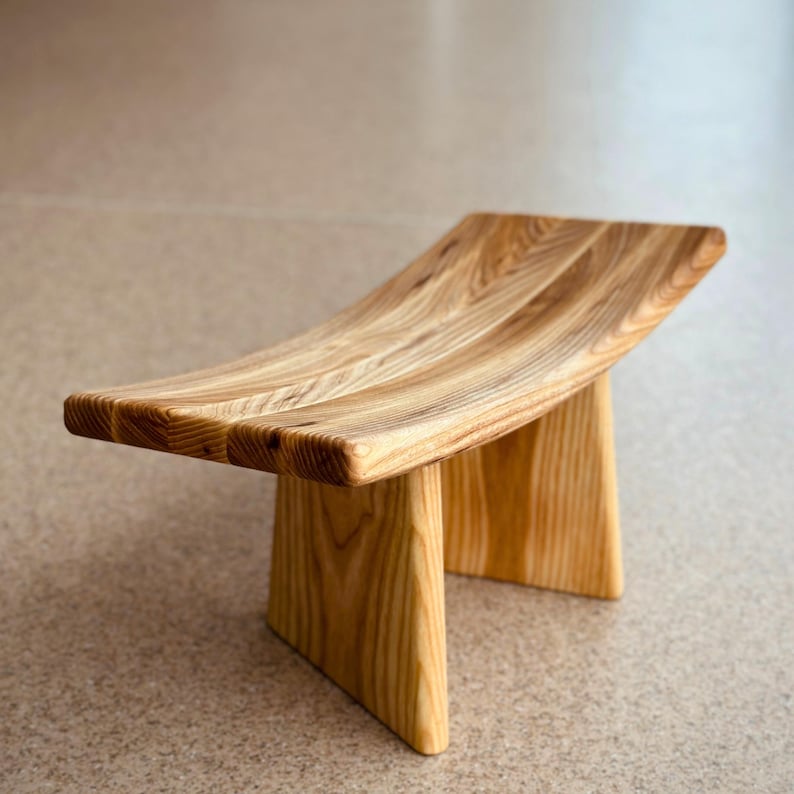 May include: A wooden meditation stool with a curved top and two legs. The stool is made of light brown wood with a natural finish.