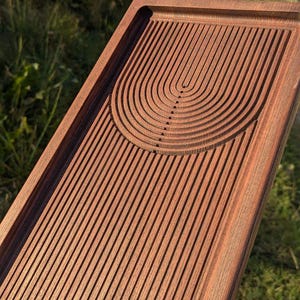 May include: A rectangular wooden tea tray with a carved design. The tray features parallel grooves across the surface, with a semi-circular pattern at one end. The wood has a reddish-brown hue, and the tray is held by a hand.