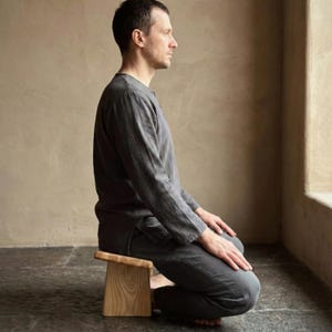 May include: A light-colored wooden meditation stool with a rectangular seat and a slightly angled top. The stool is made of light-colored wood and is approximately 12 inches long. The person is wearing a gray outfit.
