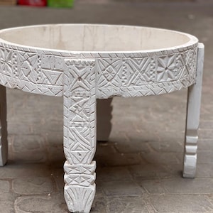 May include: White wooden round table with intricate carved designs on the top and legs. The table has a simple, rustic style.