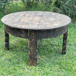 May include: A round, dark brown wooden table with carved details on the tabletop and legs. The table has a low profile and is set on a grassy surface. The table's diameter is approximately 91 cm.