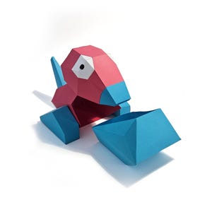 May include: A papercraft Porygon figure, with a red head and body and blue details. The figure is made of angular shapes and is set against a white background. The design includes a blue beak and tail.