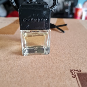 May include: A black and clear glass car air freshener with a black string hanging from the top. The air freshener has the text "Car Perfume" printed on the front.