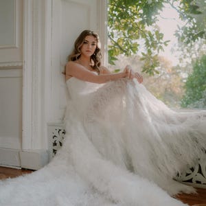 Może przedstawiać: Kobieta w białej, bez ramiączek sukni ślubnej z piórkową spódnicą siedzi przy oknie. Suknia ma przezroczysty gorset i spływającą spódnicę. Kobieta patrzy w obiektyw.