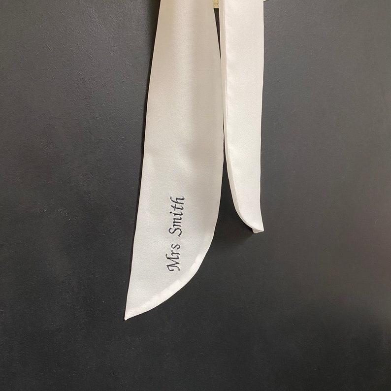 May include: White satin ribbon with the text "Mrs. Smith" embroidered in black.