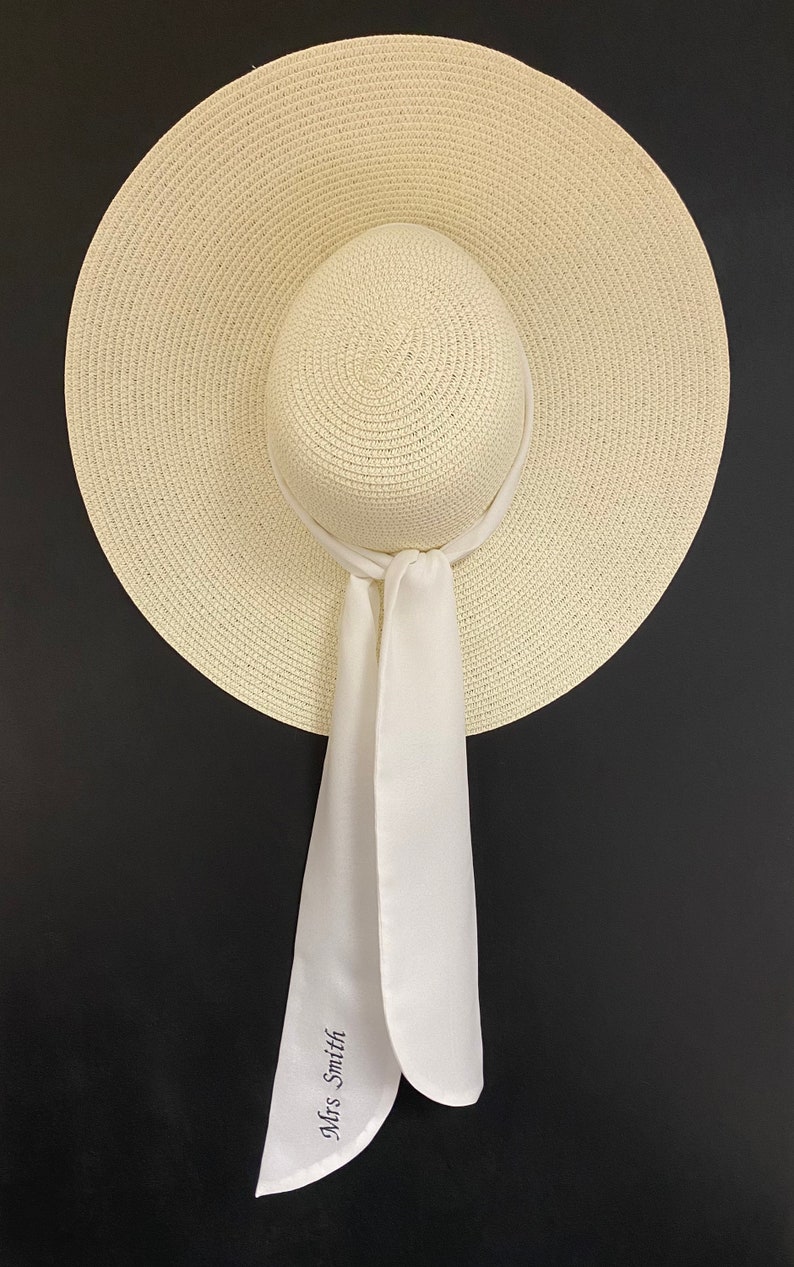 May include: A white straw sun hat with a wide brim and a white satin ribbon tied around the crown. The ribbon has the text "Mrs. Smith" embroidered on it.