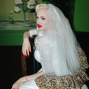 May include: A bride in a white lace wedding dress with a veil and a leopard print trim. The bride has blonde hair, red lipstick, and red nail polish. The background features a green wall and a floral mural.