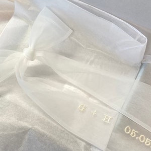 May include: White sheer ribbon with a bow tied in the center. The ribbon has the initials "G + H" and the date "05.05.20" embroidered in gold thread.