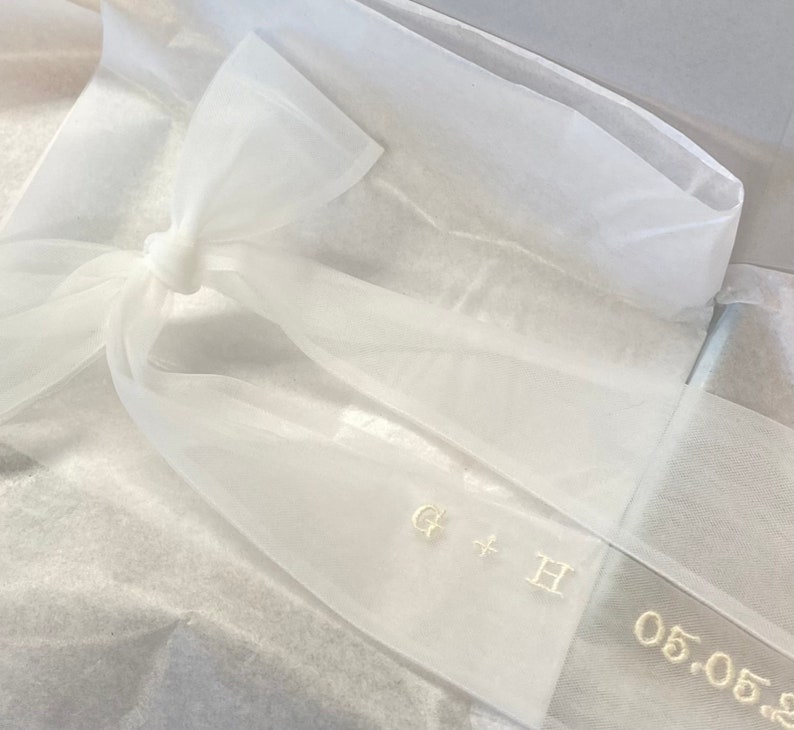May include: White sheer ribbon with a bow tied in the center. The ribbon has the initials "G + H" and the date "05.05.20" embroidered in gold thread.