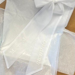 May include: White sheer ribbon with a bow tied at the top. The ribbon has the text "M + M" and "08.18.23" embroidered on it.