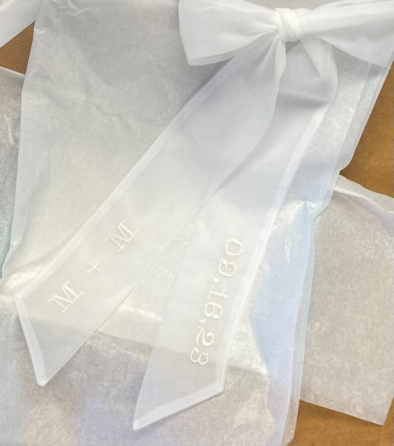 May include: White sheer ribbon with a bow tied at the top. The ribbon has the text "M + M" and "08.18.23" embroidered on it.