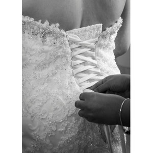 May include: A white wedding dress with a lace bodice and a corset back. The corset is being tied by a person's hands.