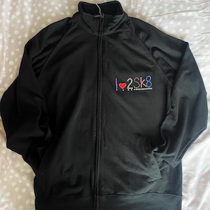 以下が含まれることがあります： 黒色のジップアップトラックジャケット。ハイカラーとサイドポケット付き。左胸にはカラフルなラインストーンで「I ❤️ 2SK8」の文字が施されています。金属製のハンガーにかかっています。