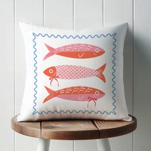 May include: A white square throw pillow with a blue wavy border. The pillow features three stylized fish in shades of orange and pink. The pillow is resting on a small wooden stool with white legs.