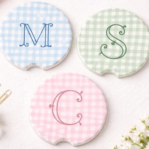 May include: Three round car coasters with a gingham pattern in blue, green, and pink. Each coaster features a different initial: "M", "S", and "C" in coordinating colors. The coasters are made of ceramic.