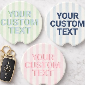 May include: Three striped car coasters in pastel colors: green, blue, and pink. Each coaster has a semi-circular notch and the text "YOUR CUSTOM TEXT" in a matching color. A car key with a gold key ring and a black key fob is next to the coasters.