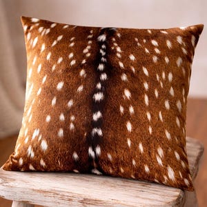 May include: A square throw pillow featuring a textured, faux-fur finish with a brown base color, white spots, and a dark brown central stripe running vertically down the middle, mimicking the appearance of a deer hide pattern.