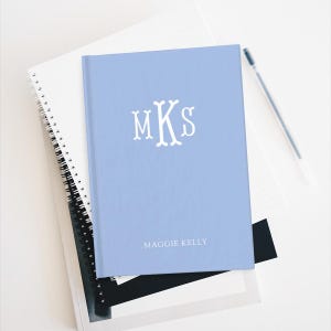 May include: A light blue hardcover notebook with the initials "MKS" in white. The name "Maggie Kelly" is printed below the initials. The notebook is stacked on top of two other notebooks and a pen.