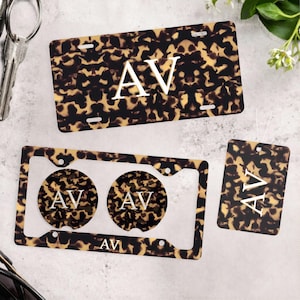 May include: A rectangular license plate, a rectangular license plate frame with two circular cup holder inserts, and a rectangular air freshener, all featuring a brown and black tortoiseshell pattern and the white monogram letters AV.