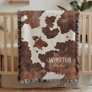 May include: A brown and white cowhide-patterned baby blanket with the name "WINSTON blake" printed in white. The blanket has a fringed edge and is draped over a wooden crib.