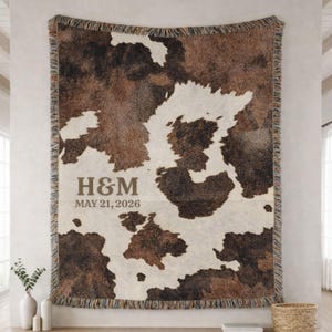 May include: A woven throw blanket featuring a brown and white cowhide pattern with fringed edges. The blanket displays the text H&M and the date MAY 21, 2026, printed in a brown serif font on the lower left side of the patterned fabric surface.
