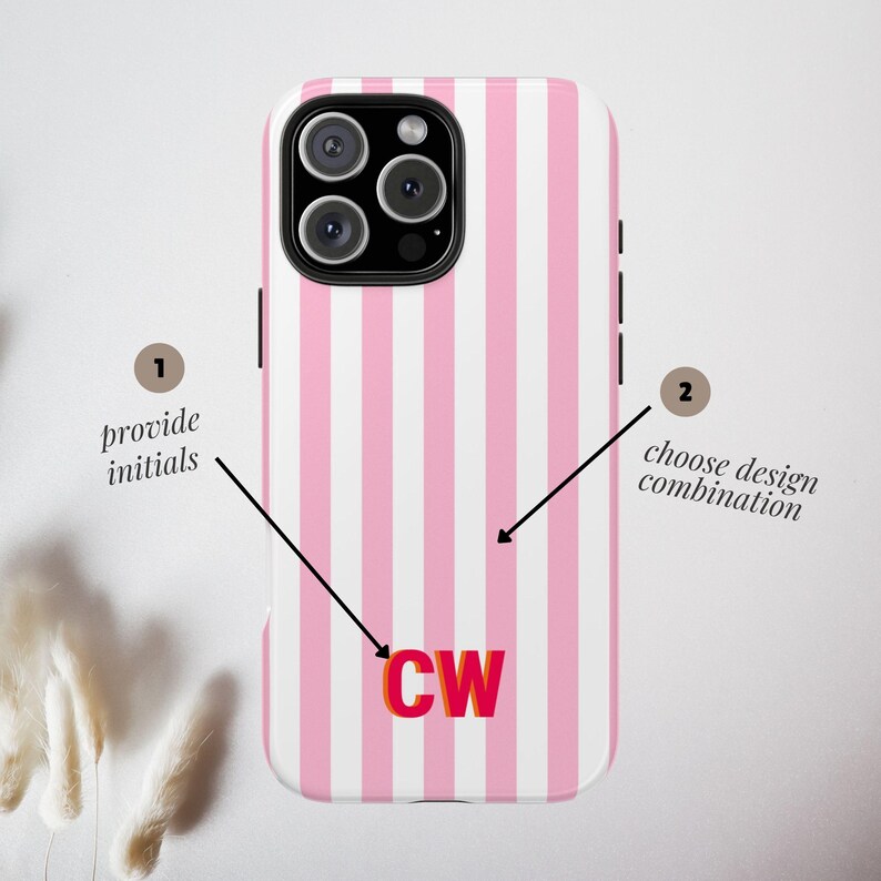 May include: A smartphone case featuring a vertical pink and white striped pattern. The center displays the initials CW in bold red and orange lettering. Two black arrows point to the case, with text labels reading 1 provide initials and 2 choose design combination.