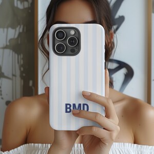 May include: A smartphone case featuring a vertical light blue and white striped pattern. The case has a dark gray camera module cutout at the top left and the letters BMD printed in a bold, dark blue sans-serif font in the lower center area.