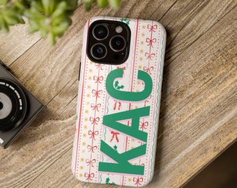 May include: A white smartphone case featuring a vertical pattern of red bows, green holly leaves, and gold stars. The letters KAC are printed in large, bold green font vertically down the center of the case, which has a triple camera lens.