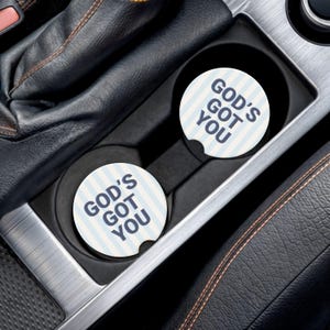 May include: Two white car coasters with blue and white vertical stripes and the text "GOD'S GOT YOU" in navy blue. The coasters are in a car's cup holders, with a black interior and silver trim.