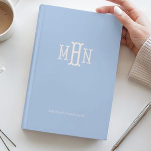 May include: A light blue journal with the initials "MHN" in a decorative font. The name "Minnah Harrison" is printed below the initials. A silver pen and a cup of coffee are in the background.