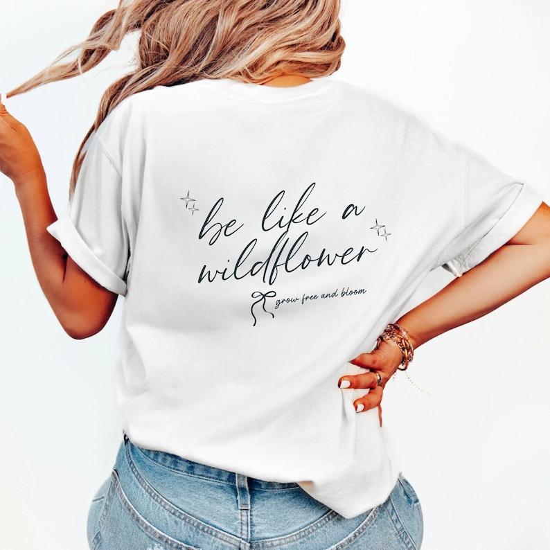 K&ouml;nnte beinhalten: Ein wei&szlig;es kurz&auml;rmeliges T-Shirt mit schwarzem Text auf der R&uuml;ckseite, der in Schreibschrift "be like a wildflower" lautet, begleitet von kleinen Sternsymbolen und einer Schleifengrafik &uuml;ber dem Satz "grow free and bloom" in kleinerer Schrift.