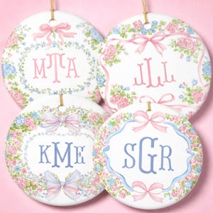 May include: Four round white ceramic ornaments featuring floral wreaths of pink and blue hydrangeas with pink or blue ribbons. Each ornament displays a unique three-letter monogram in either pink or blue serif font, suspended by gold strings.