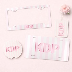 May include: A set of items featuring a light pink and white vertical striped pattern with the letters KDP printed in pink. The set includes a rectangular license plate frame, a rectangular license plate, a circular coaster, and a rectangular tag.