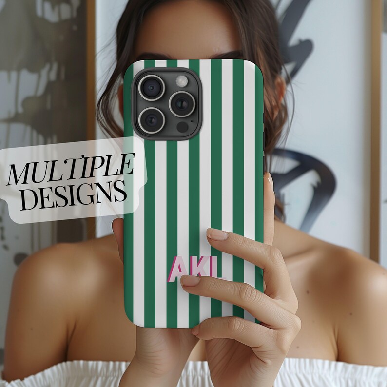 May include: A phone case with a green and white striped pattern. The case has the letters "AKI" in pink. The phone case is held in front of a person. The text "MULTIPLE DESIGNS" is in a white banner.