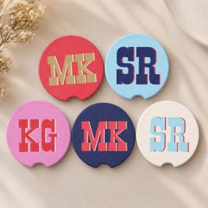 May include: Five circular coasters with a small notch on the bottom edge. The top left is red with gold MK letters, top right is light blue with navy SR letters, bottom left is pink with red KG letters, bottom center is navy with red MK letters, and bottom right is cream with light blue SR letters.