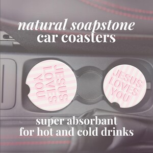 May include: Two natural soapstone car coasters with the text "JESUS LOVES YOU" in pink. The coasters are round with a striped pattern and are designed to absorb moisture from hot and cold drinks. The image also includes the text "super absorbant for hot and cold drinks".